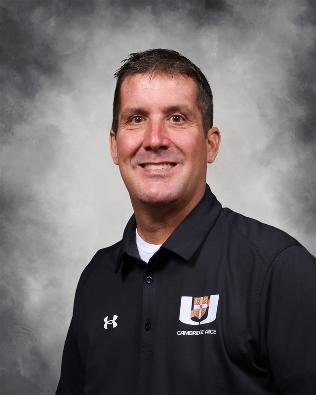 A smiling man in a black polo shirt with white text and logo, standing against a gray background.