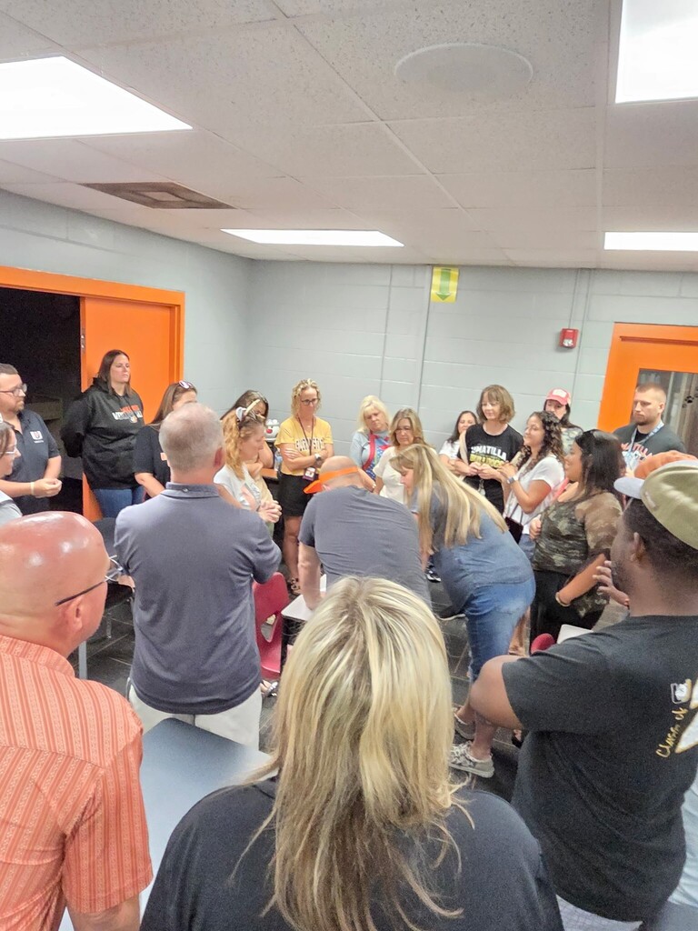 People gather in a room with bright overhead lights, some seated, others standing. Two orange doors are visible.