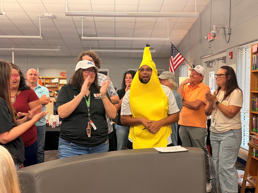 Group of people standing and clapping in a room, one person in a yellow banana costume, US flag on wall.