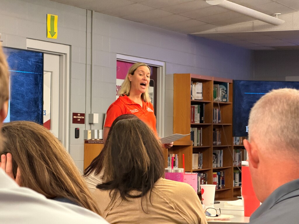 A woman in orange stands in a classroom, speaking to a group of people seated at tables.