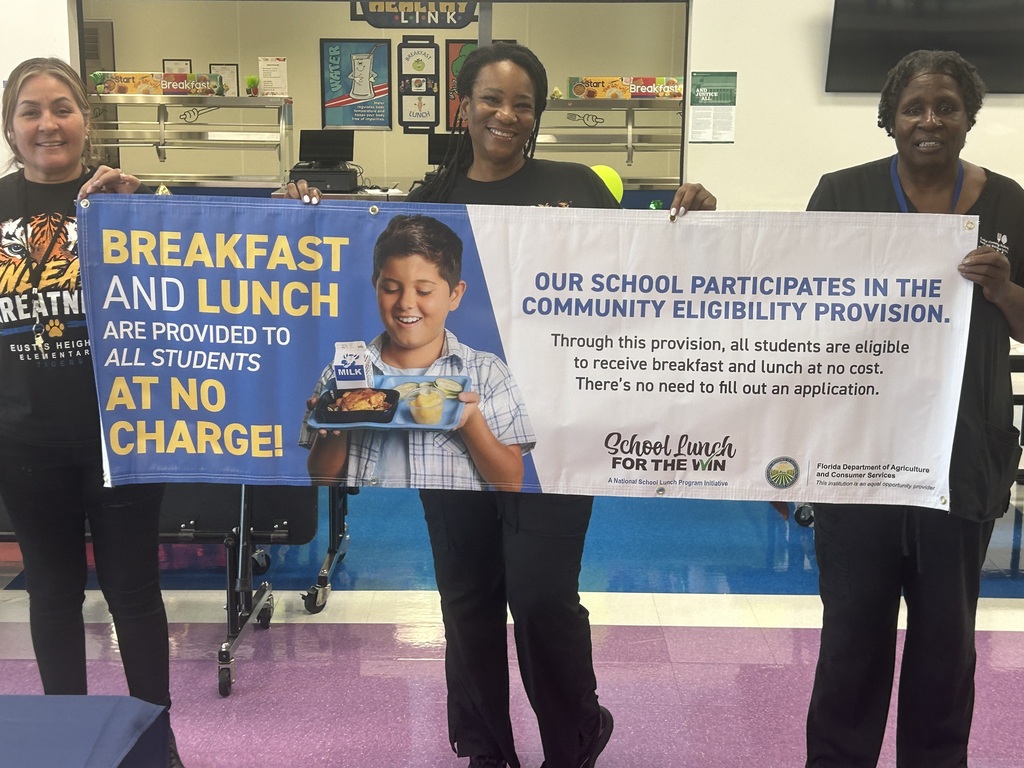 Four women hold a sign with a child, reading "Breakfast and Lunch are provided to all students at no charge!"