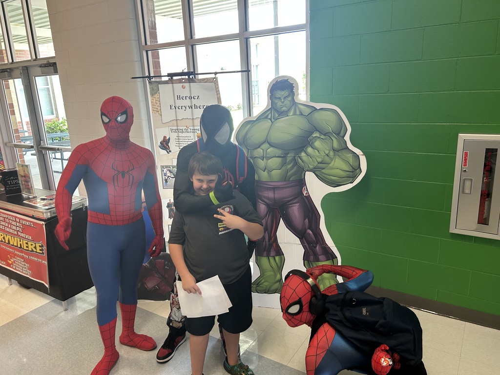 Four people stand in a hallway, two in superhero costumes, one holding a paper, and another crouching. Behind them, a green wall with cutouts of superheroes.