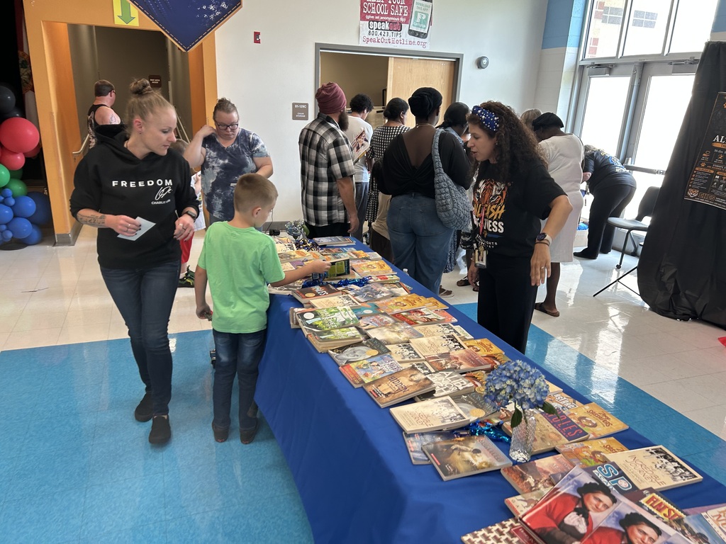 A child at a long table of books, with two women looking at them. Behind them, several adults are standing and talking.