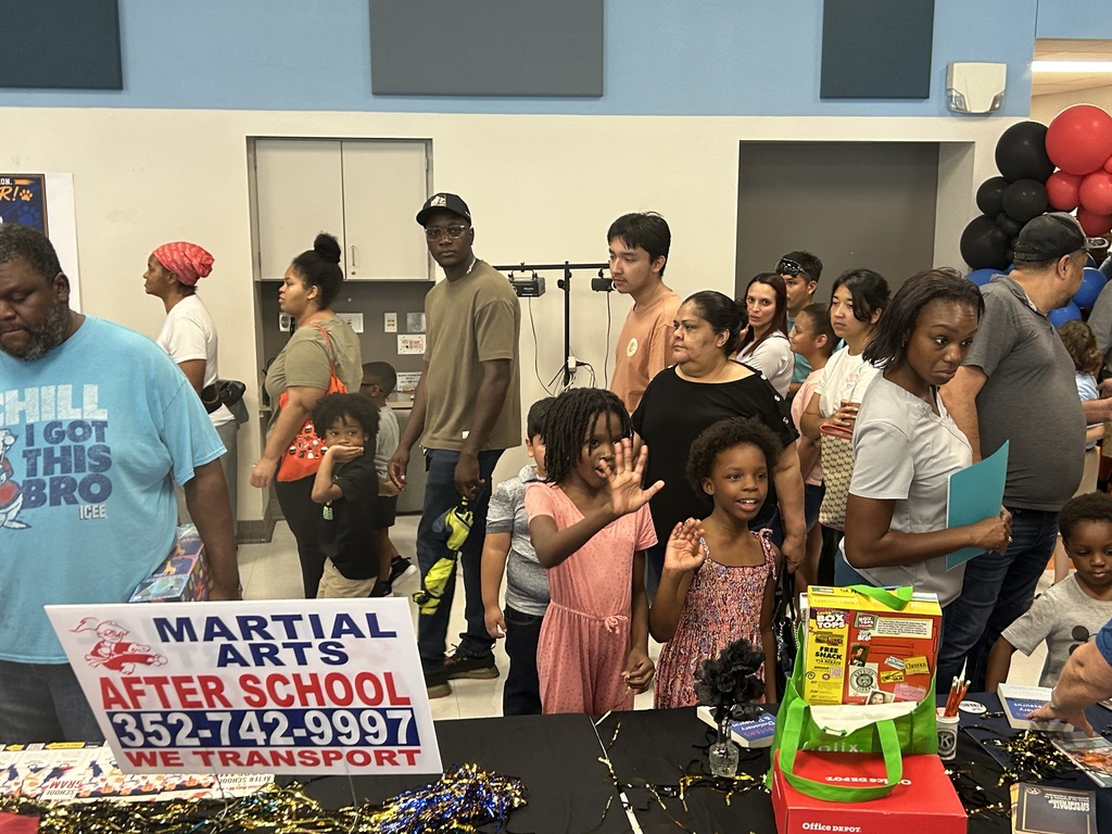 A crowd of people, some wearing hats, stands around a table with a sign that reads "Martial Arts After School."