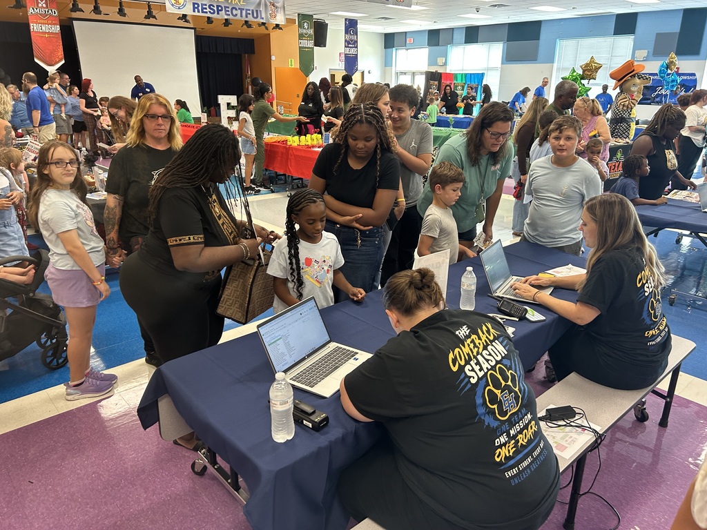 People sit at tables in a classroom with laptops; one adult in black speaks to a child.
