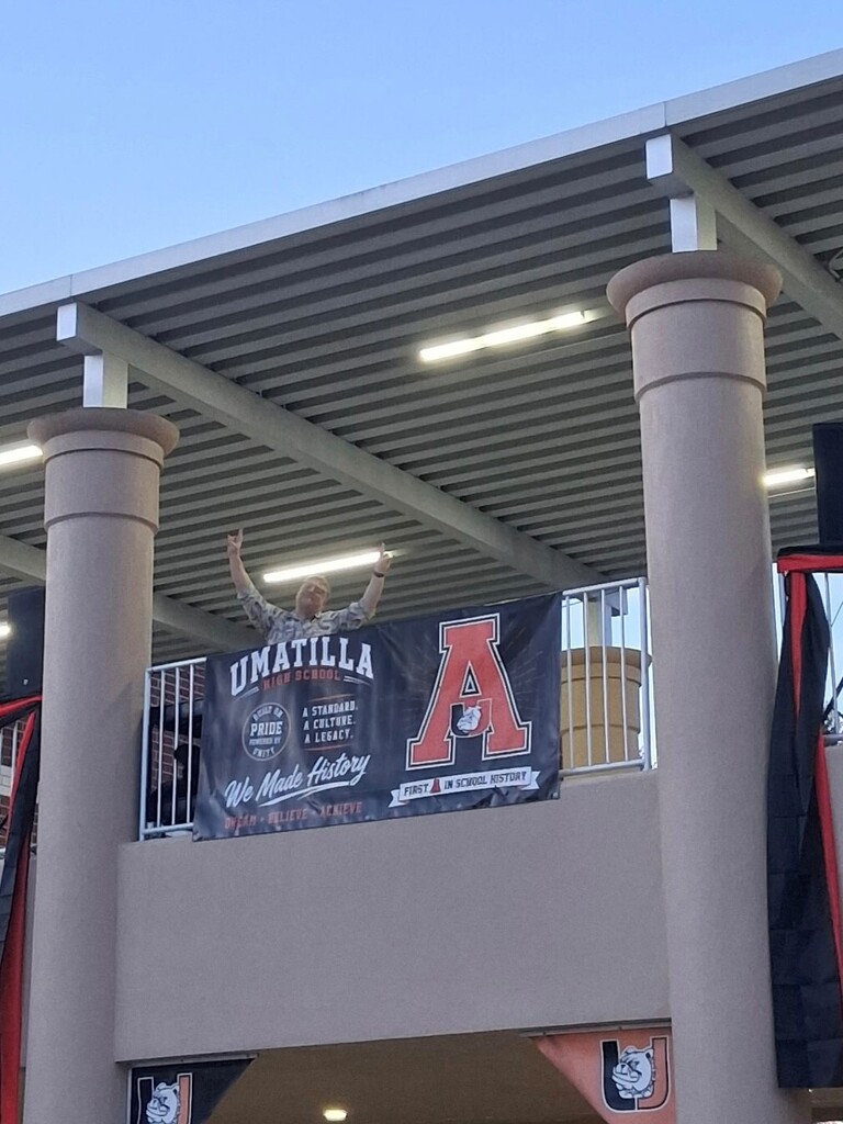 A person raising their hands stands on a balcony with a banner reading "UMATILLA A". The banner is held by a railing.