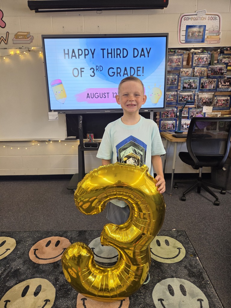 A young boy stands in a classroom holding a large gold number three balloon, with a screen in the background displaying a celebratory message.