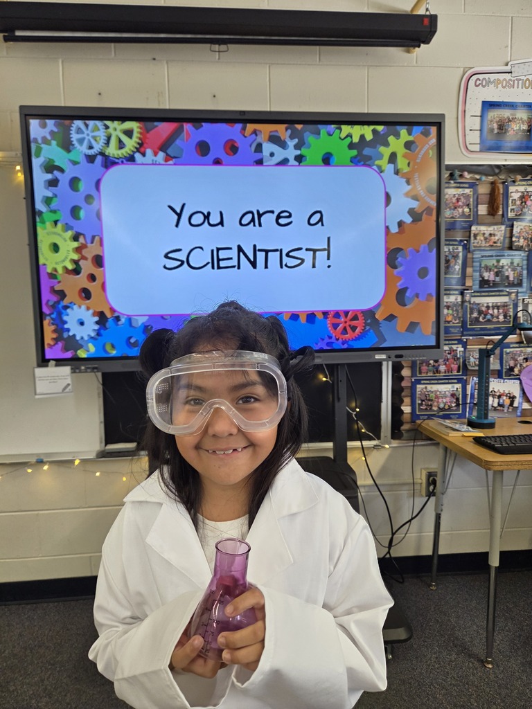 A young girl in a white lab coat and goggles stands in front of a screen. She holds a purple flask.
