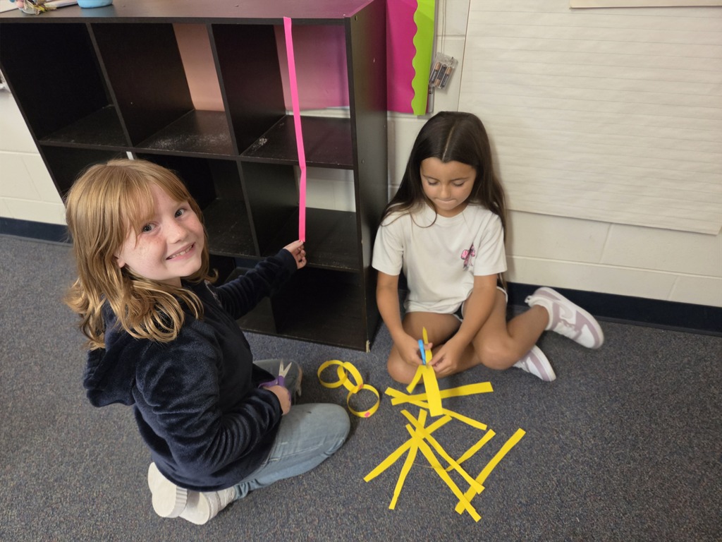 Two children sitting on floor, one holding pink rod, other using scissors, surrounded by yellow cut strips.