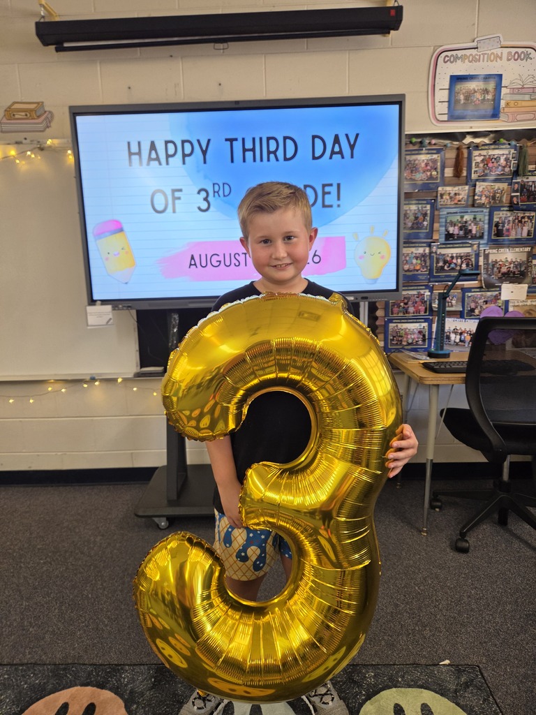 A boy holding a gold foil balloon shaped like the number 3, with a monitor displaying a message behind him.