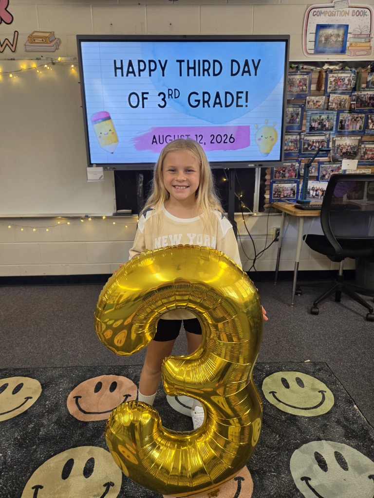 A young girl stands with a large, golden number three balloon in a classroom with a smiley-face rug.