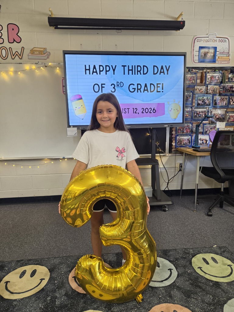 A young girl holding a large gold number three balloon stands in a classroom. Behind her is a TV screen with a celebratory message.