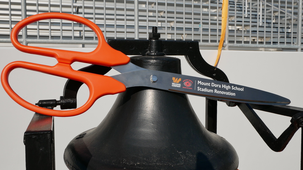 A pair of orange scissors placed over a bell with "Moses Day High School Stadium Renovation" printed. Behind, a metal fence and a white wall.