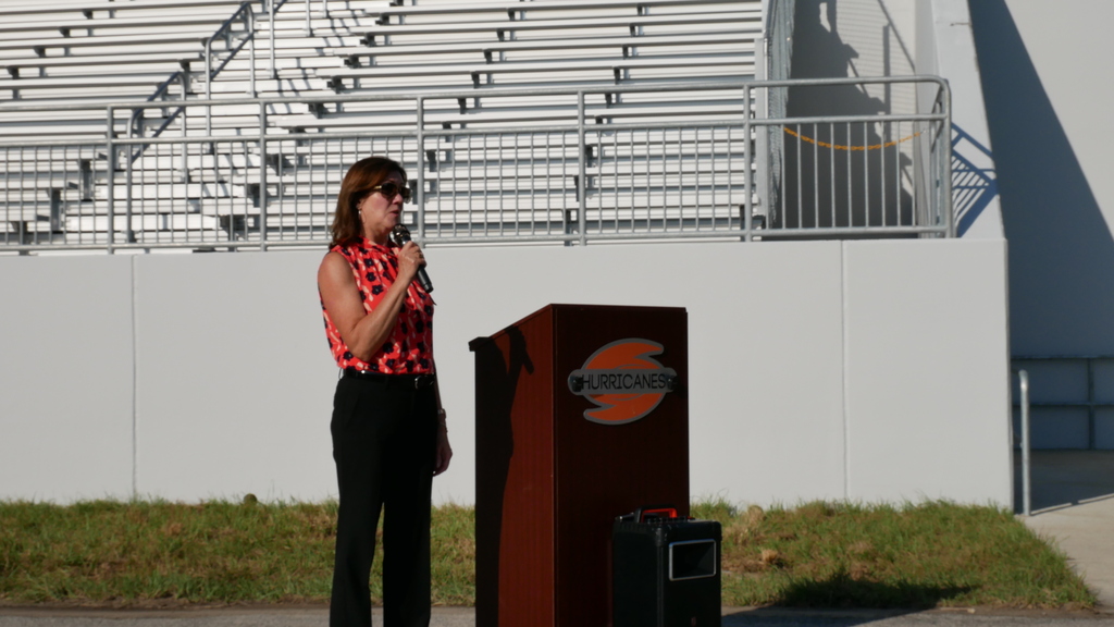 Superintendent Kornegay standing at podium in black pants and orange and black top, holding a microphone. Podium has a Hurricanes logo in orange.