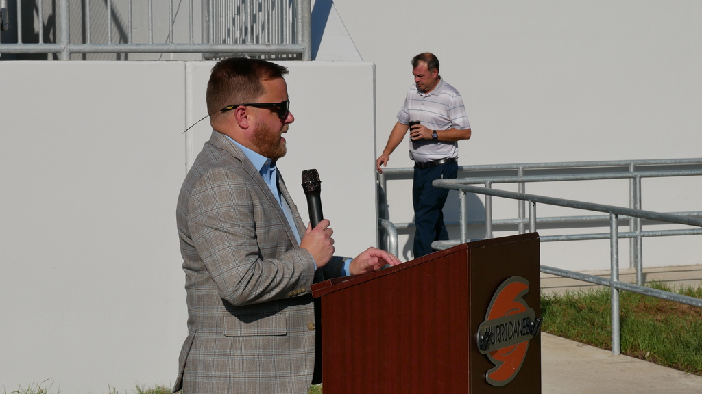 Man with microphone at podium with logo, another man standing behind a railing, white wall in background.
