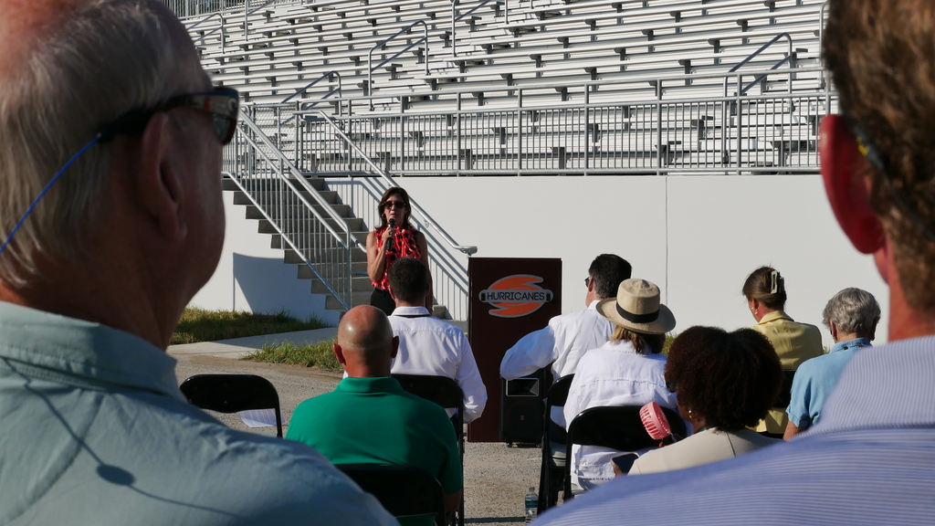 A group of people sit in chairs around a podium in a stadium. A woman speaks into a microphone.
