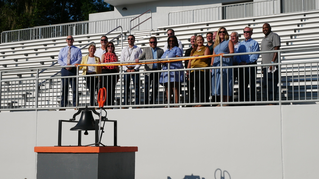 People standing in front of a railing, holding a ribbon, with a bell in front. Background includes bleachers.