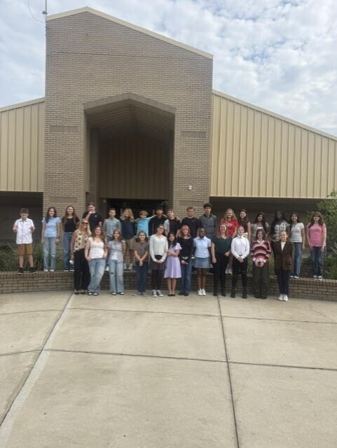 A group of students stands in front of a school building with brick walls and a large archway.