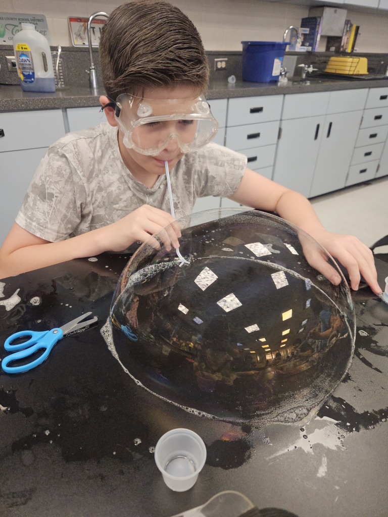 A child in a lab coat conducts an experiment with a large bubble. Behind, cabinets and a sink are visible.