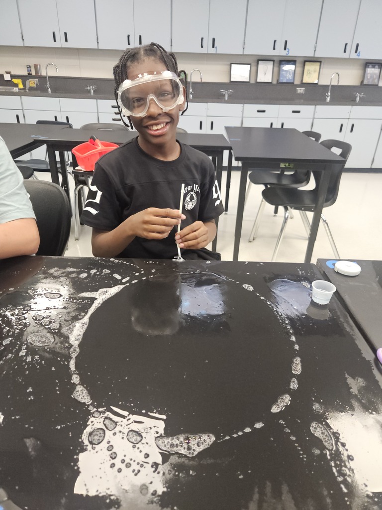 A child sits at a table with a black surface, wearing goggles and holding a pipette.
