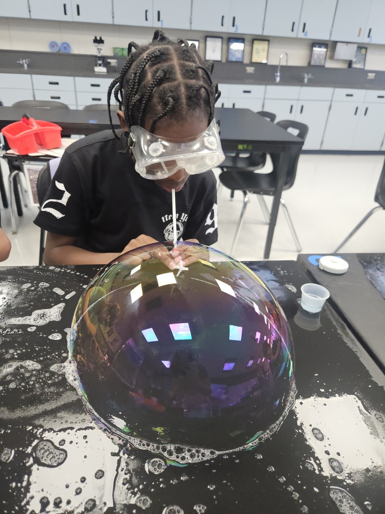 A person sits at a table in a classroom, wearing goggles and blowing a large bubble.