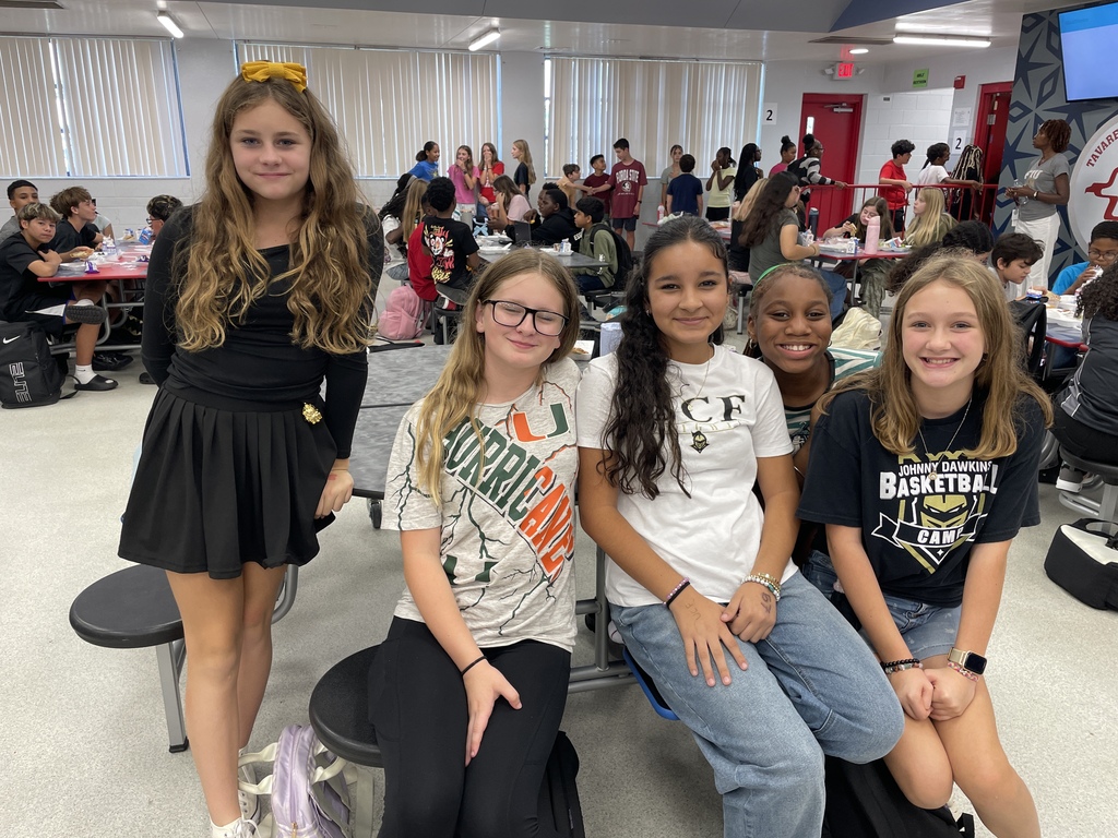 Four girls standing together in a crowded cafeteria. One girl has a flower crown on.