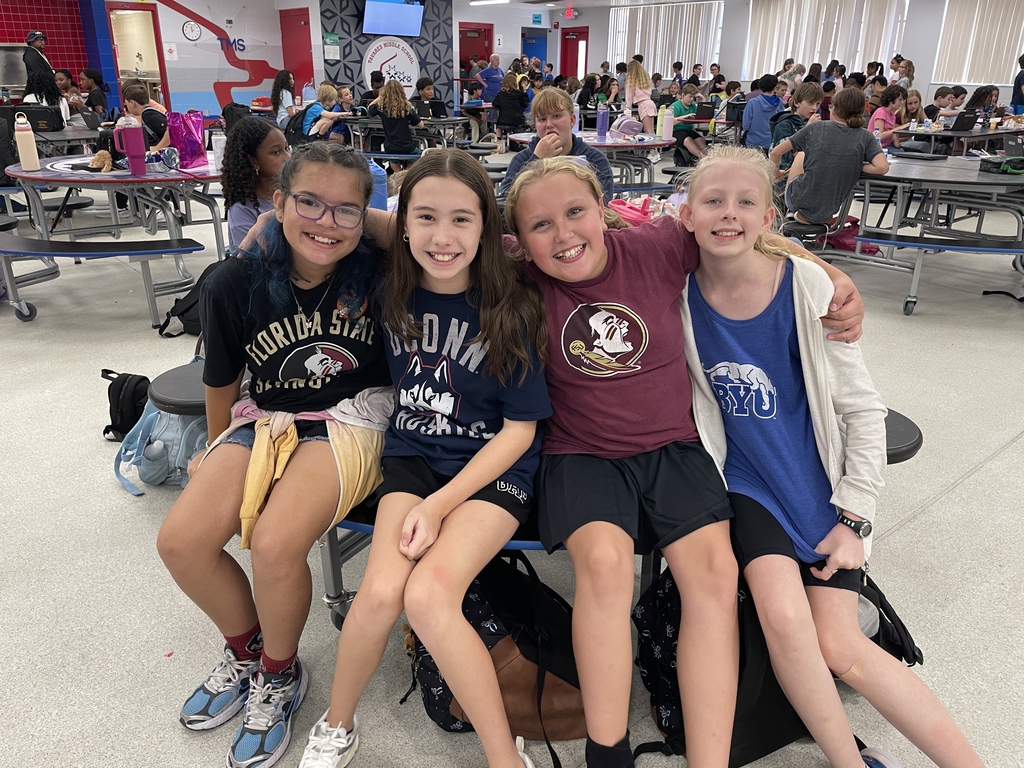 Three girls sit together in a cafeteria, smiling and posing for a photo. Two wear black, one in purple. Backpacks are nearby.