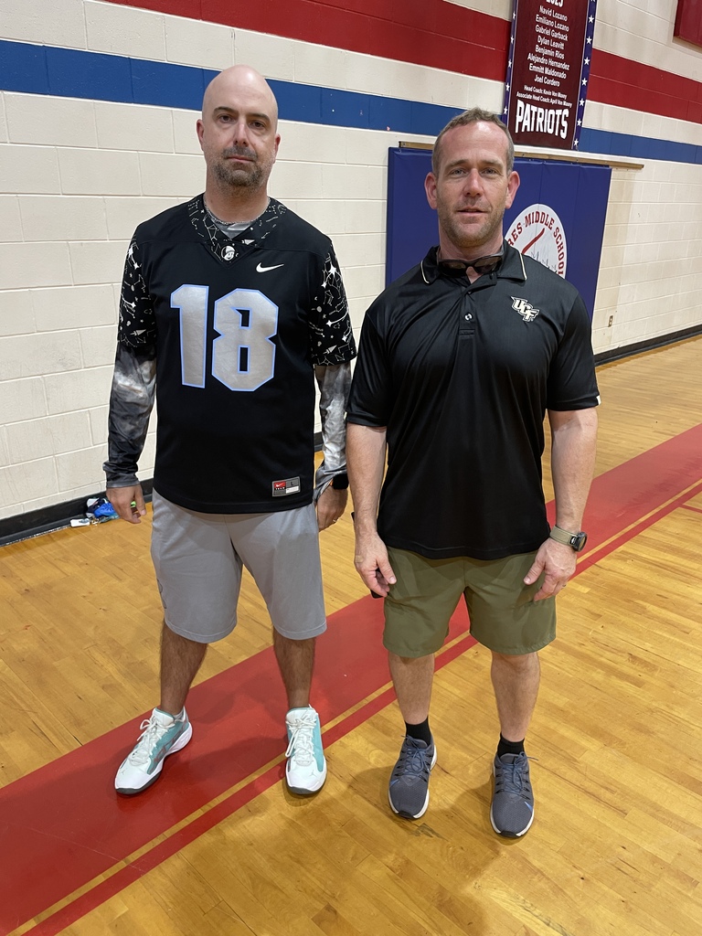 Two men on a basketball court. One wears a black jersey with the number 18 and shorts. The other wears a black polo and shorts.