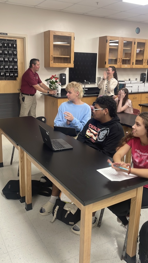 People in a classroom, seated at a table with a laptop. A person stands near a cabinet with a monitor and flowers.