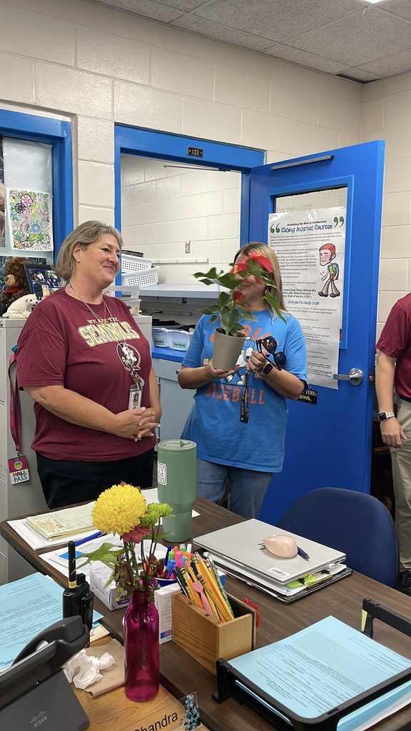 A woman with a plant on her head stands near a desk with flowers. Another woman stands nearby.