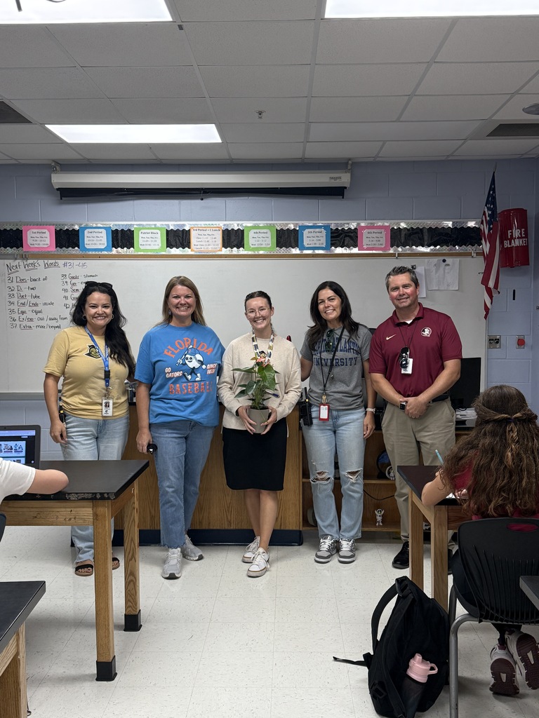 Group of people in a classroom. One person holding a potted plant. Desks, chairs, whiteboard, and flag in the background.