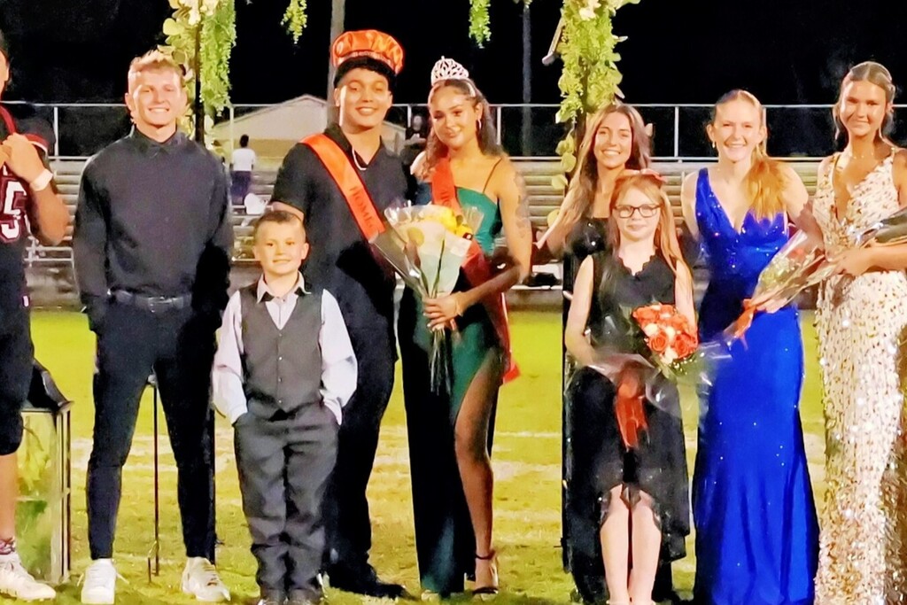 A group of individuals stands on a field, holding flowers, and wearing various attire. Some have sashes.