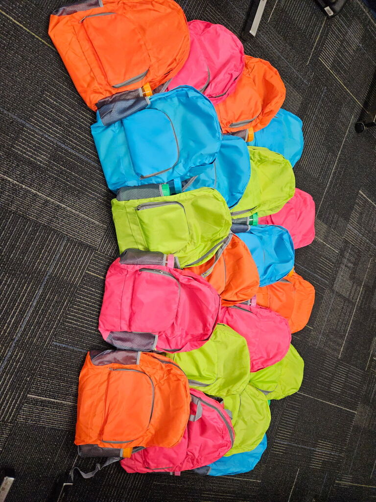 A line of backpacks in vibrant colors like orange, pink, blue, and green on a carpeted floor.