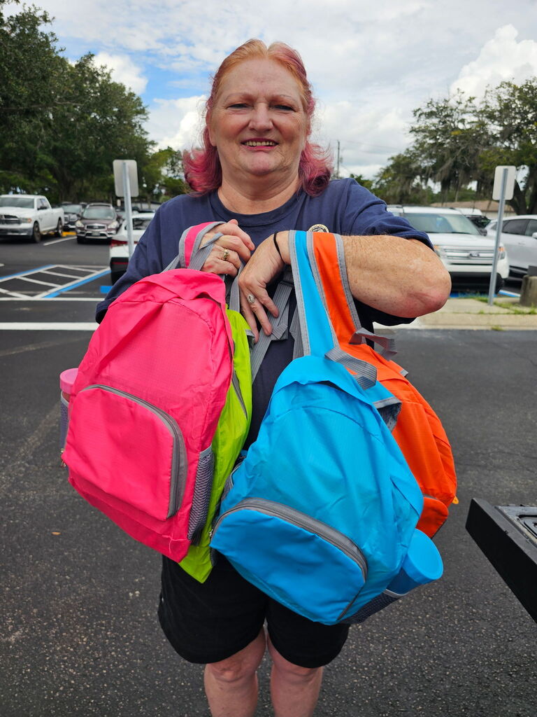 Woman standing in parking lot, holding three brightly colored backpacks, with several cars parked behind her.