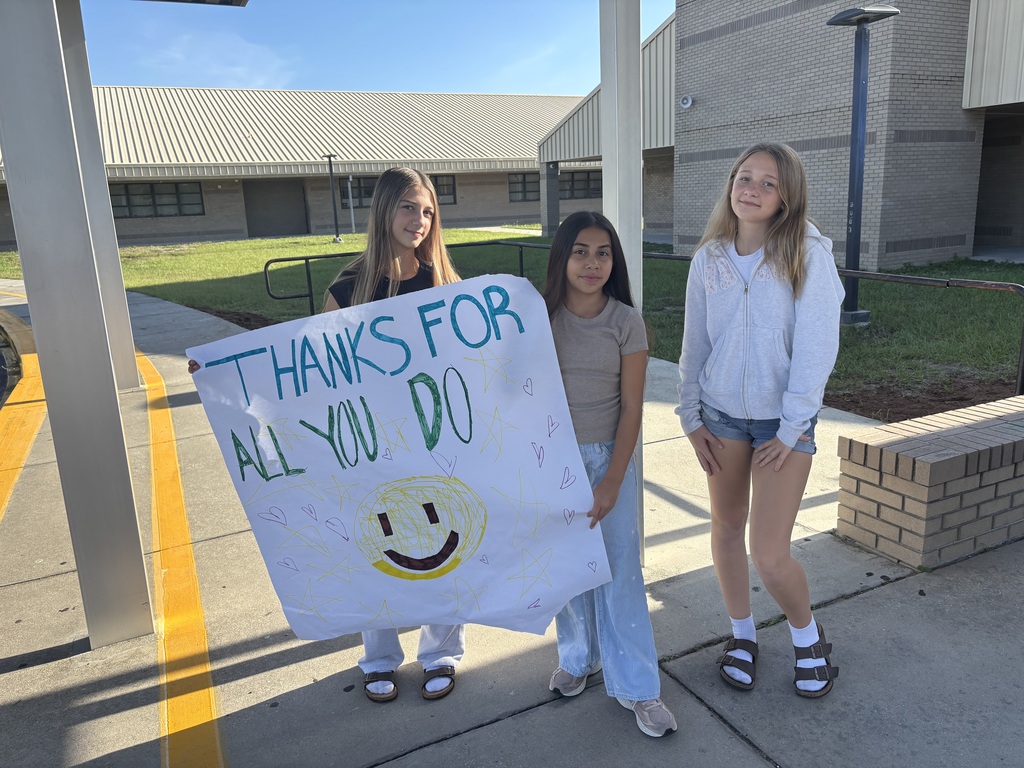 Three girls standing on a sidewalk hold a banner with a smiley face and "Thanks for all you do".