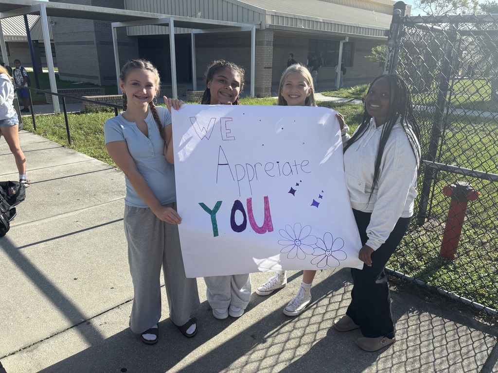 Four people hold a white sign with colorful writing in front of a chain link fence.