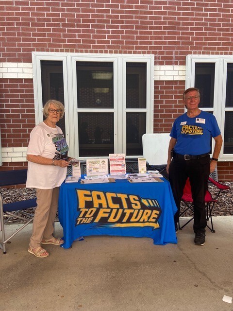 Two people stand behind a table with a blue tablecloth. The table displays a sign that reads "FACTS TO THE FUTURE".