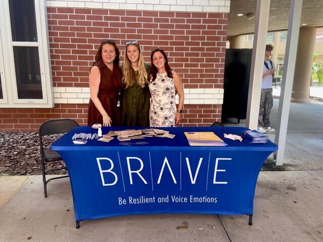 Three women stand behind a table with a blue tablecloth. The tablecloth reads "BRAVE" in white letters. A brick building is behind them.