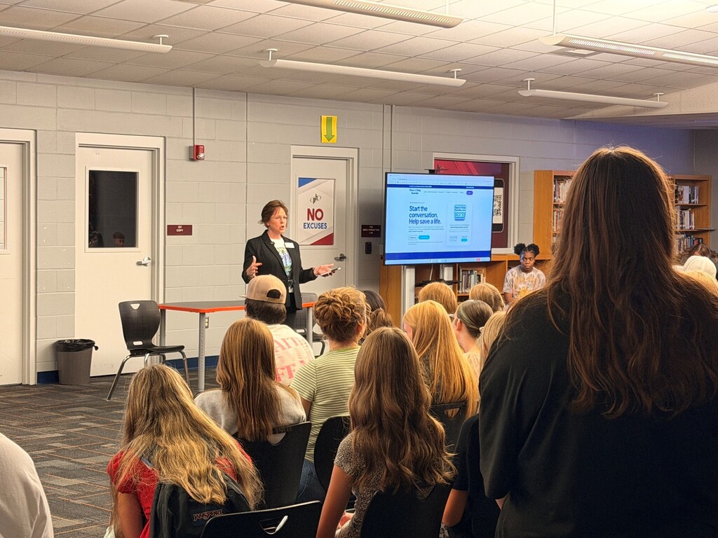 A woman stands at a podium with a screen behind her, addressing a seated audience in a room.