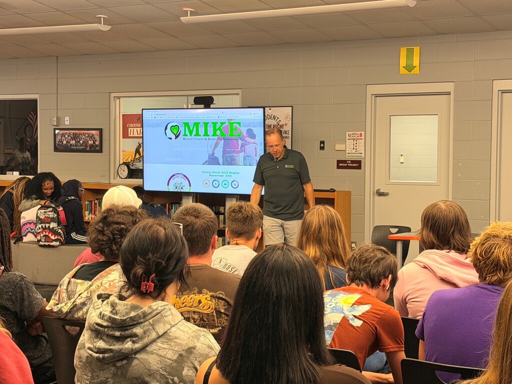 A man speaks to a seated audience in a classroom. The screen behind him shows the word "Mike."