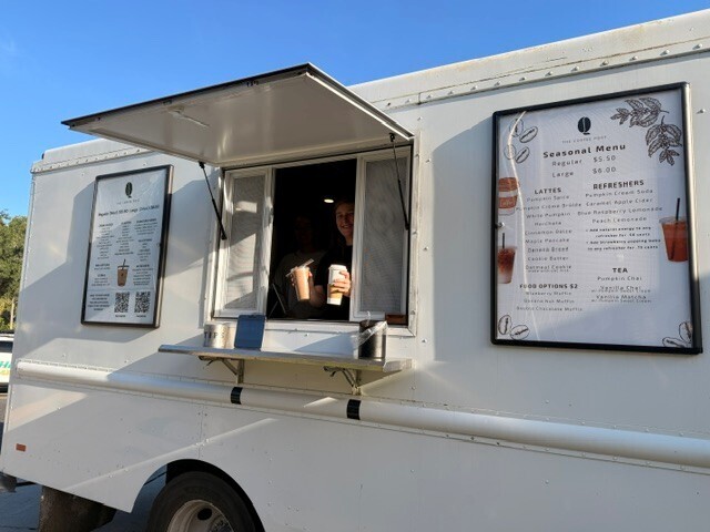 A white food truck with a menu displayed on the side. A person sits inside, holding a cup.