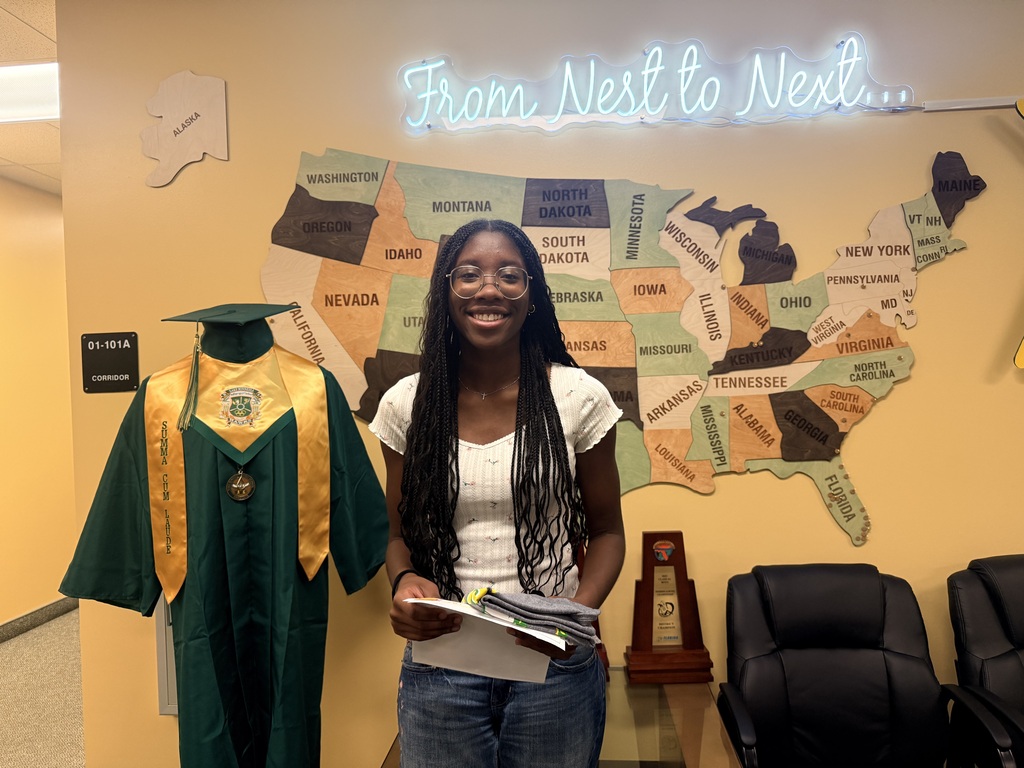 A person in a white shirt and jeans stands next to a graduation gown, holding papers. Behind them is a U.S. map on the wall and a map light with "From Nest to Next"