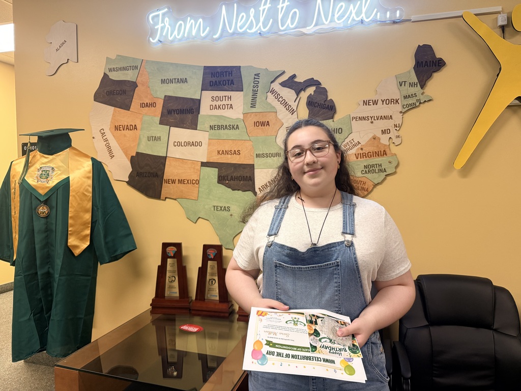 A young person stands holding a certificate in front of a U.S. map, a graduation gown, and three trophies.