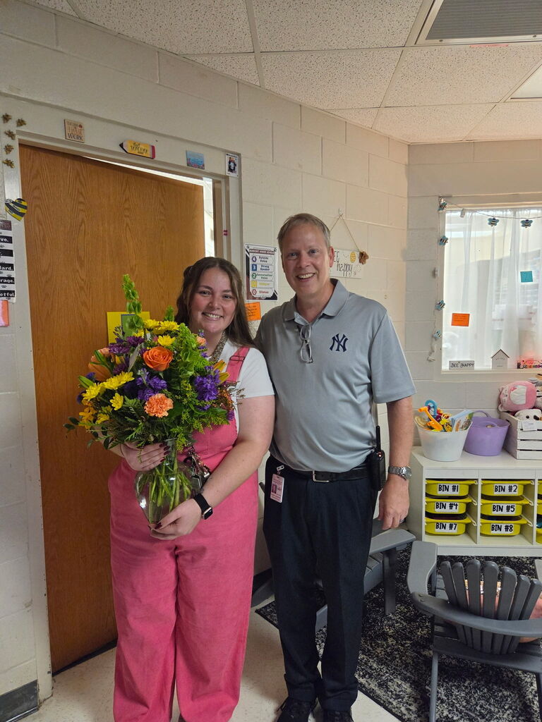 A person in pink overalls holding flowers stands next to a person in a blue shirt in a room.