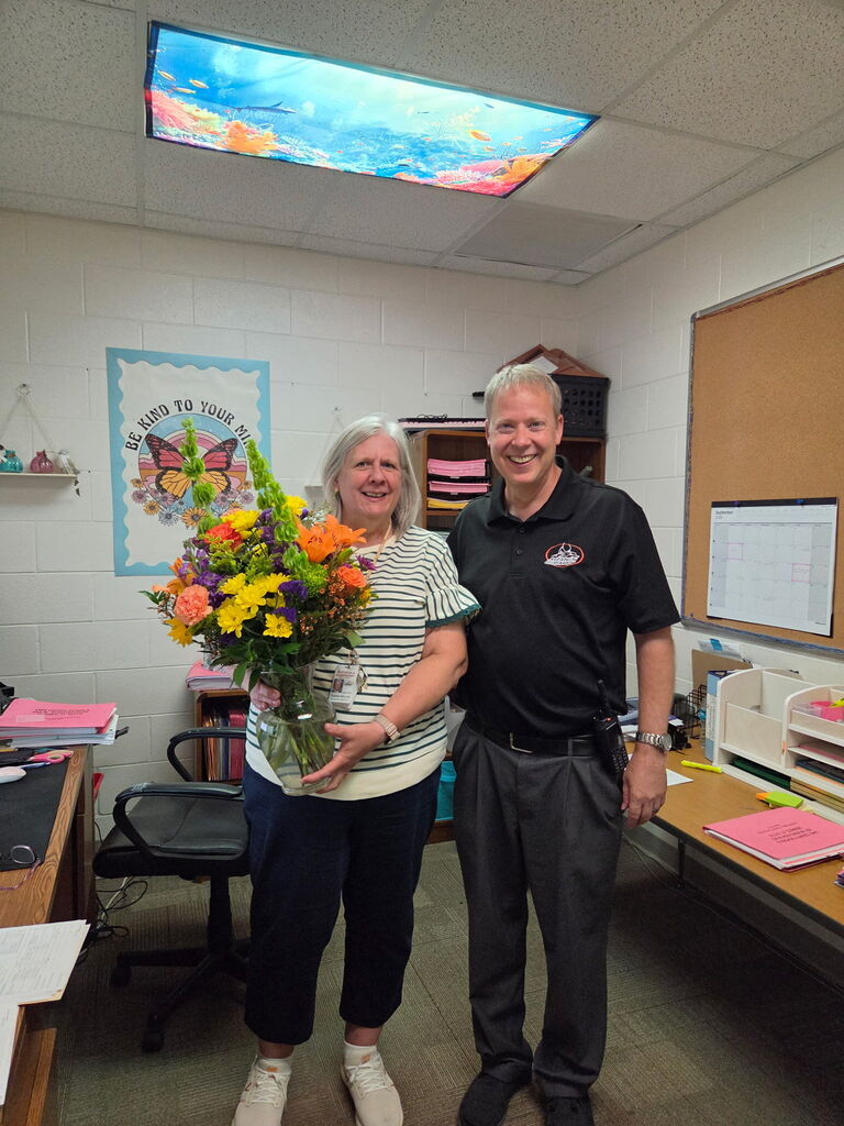 A woman holds a flower arrangement while standing next to a man in a classroom.