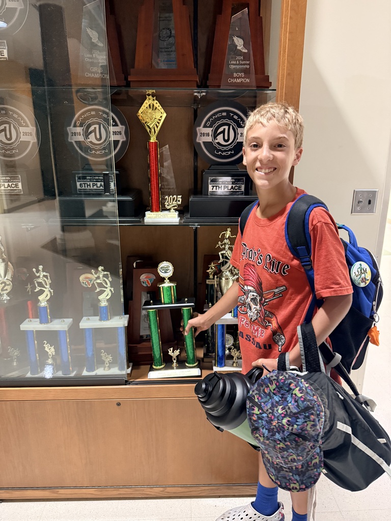 A young boy stands next to a display case with multiple trophies and a backpack in a hallway.
