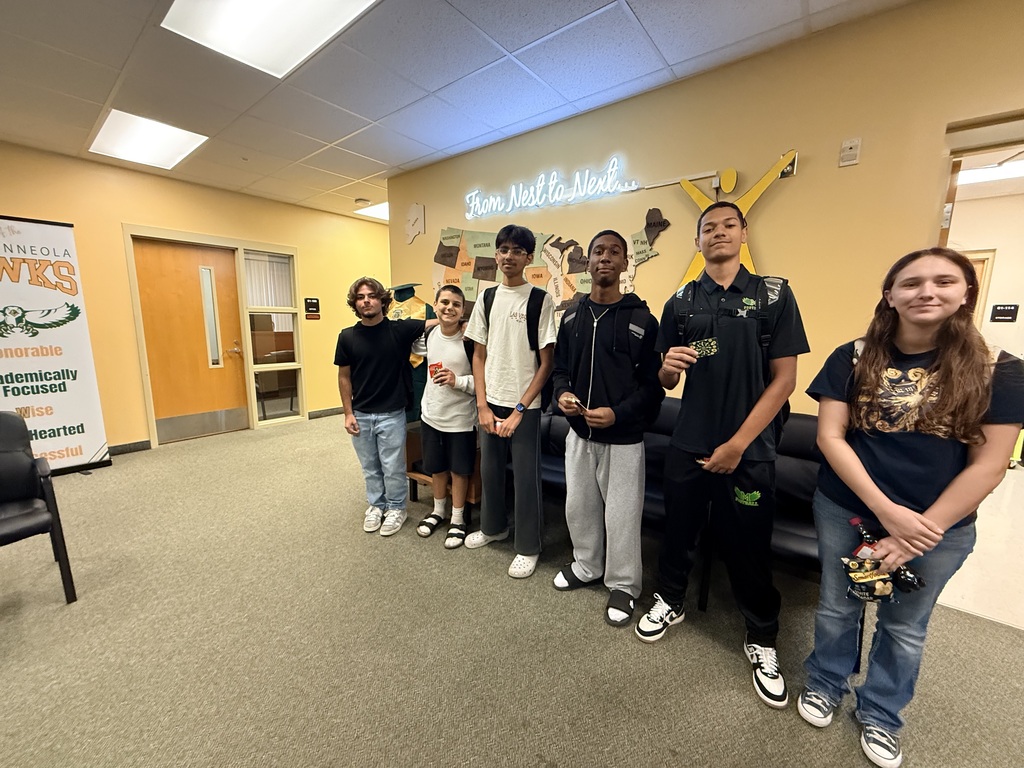 Group of students with backpacks in a hallway, smiling. Behind them is a wall with a lit sign.