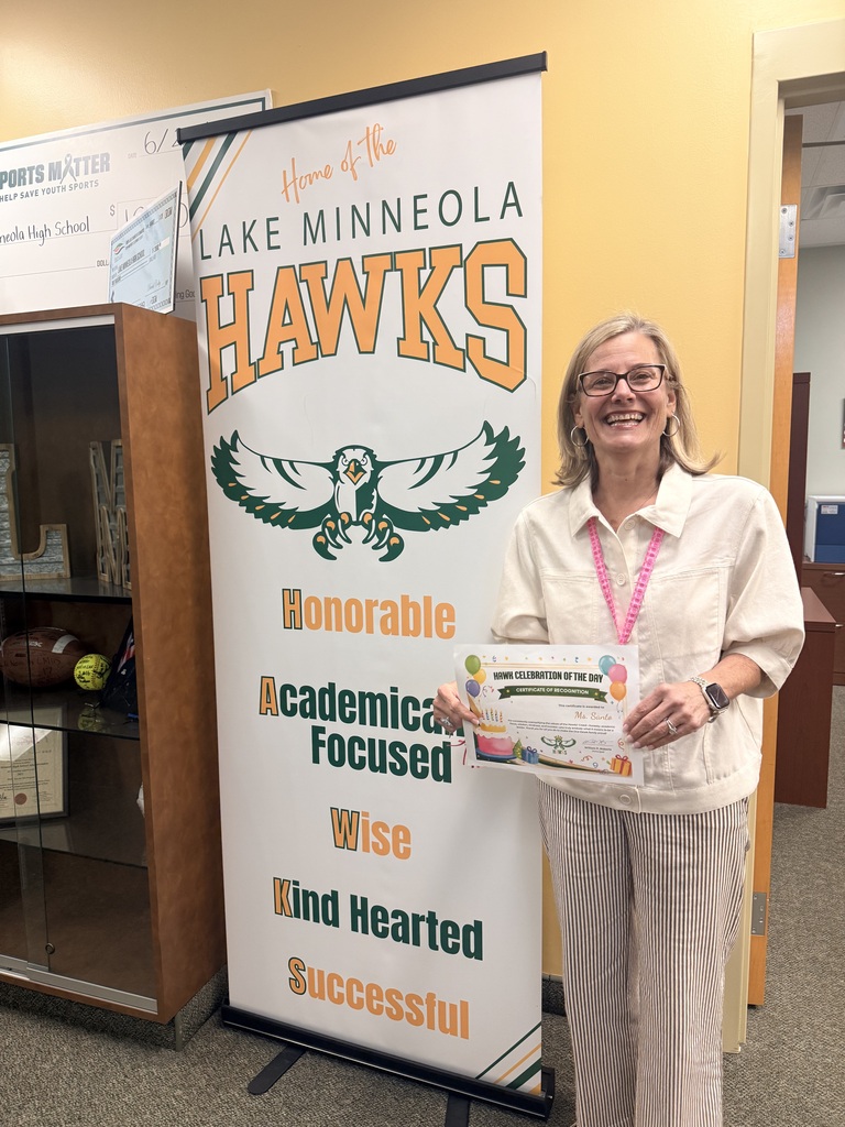 A smiling woman holds a certificate next to a banner with the words "Lake Minneola Hawks" on it.