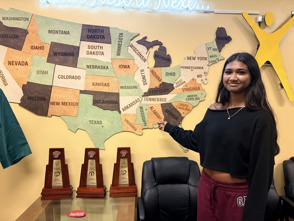 A woman stands in front of a map of the United States and three trophies on a desk.