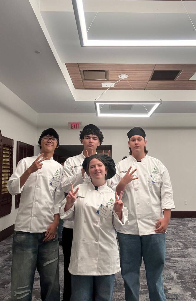 Four people in chef uniforms stand in a room, smiling and making peace signs. They have hats on.